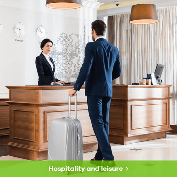 Hospitality abd Leisure - image of a suited person arriving at a hotel reception to check in. They have a md-sized wheely suitcase with them.